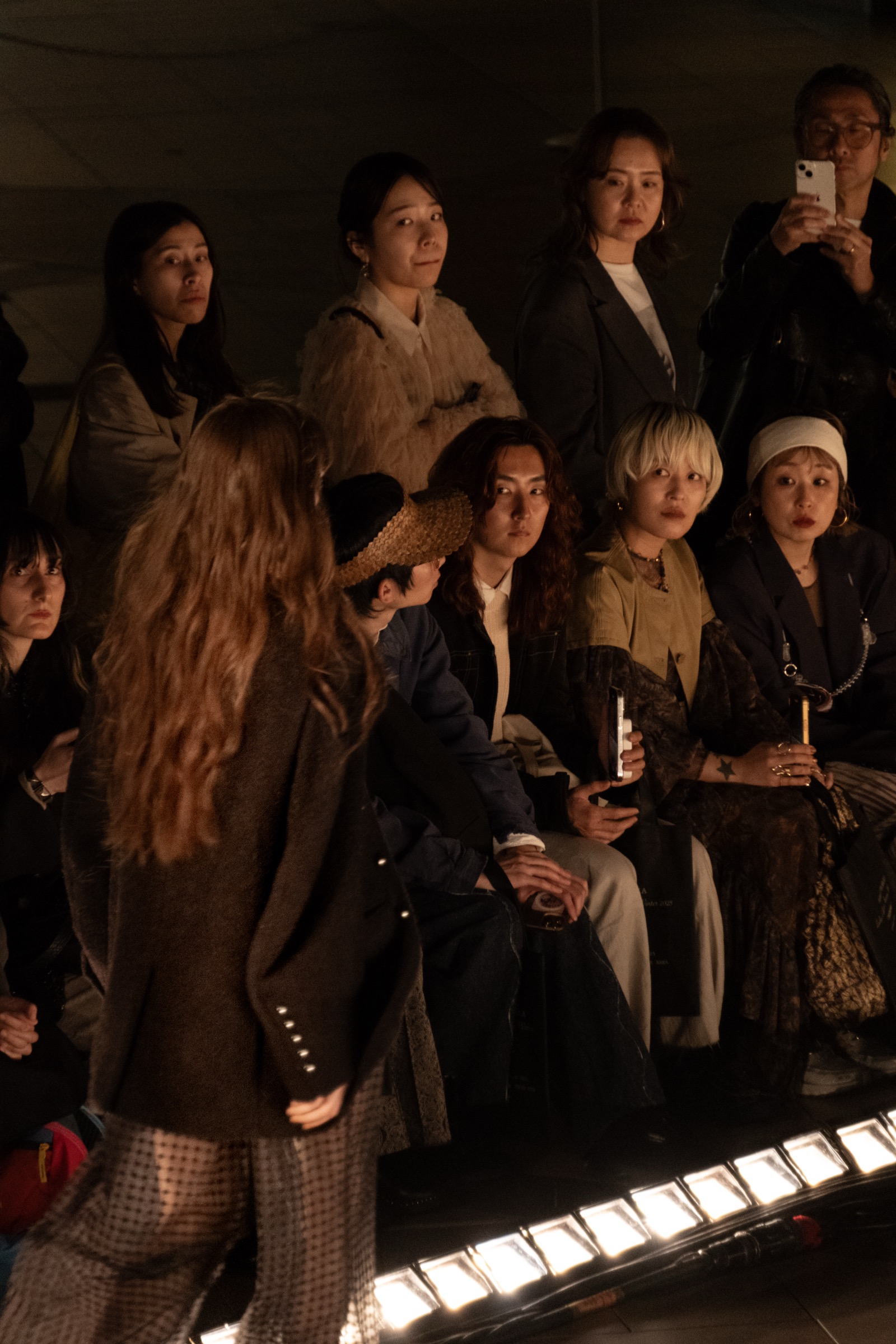 A model walks a runway as fashion-show attendees in dark coats watch from the front row, stage lights glowing below