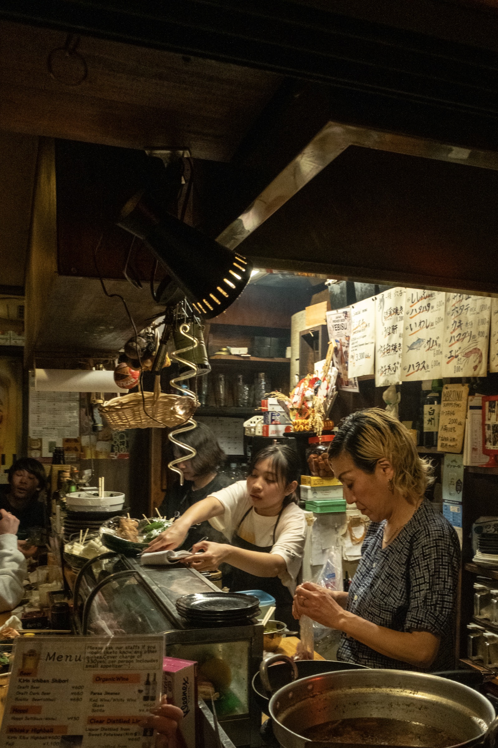 Two women work behind the counter of a tiny izakaya kitchen, handwritten menus and hanging baskets overhead