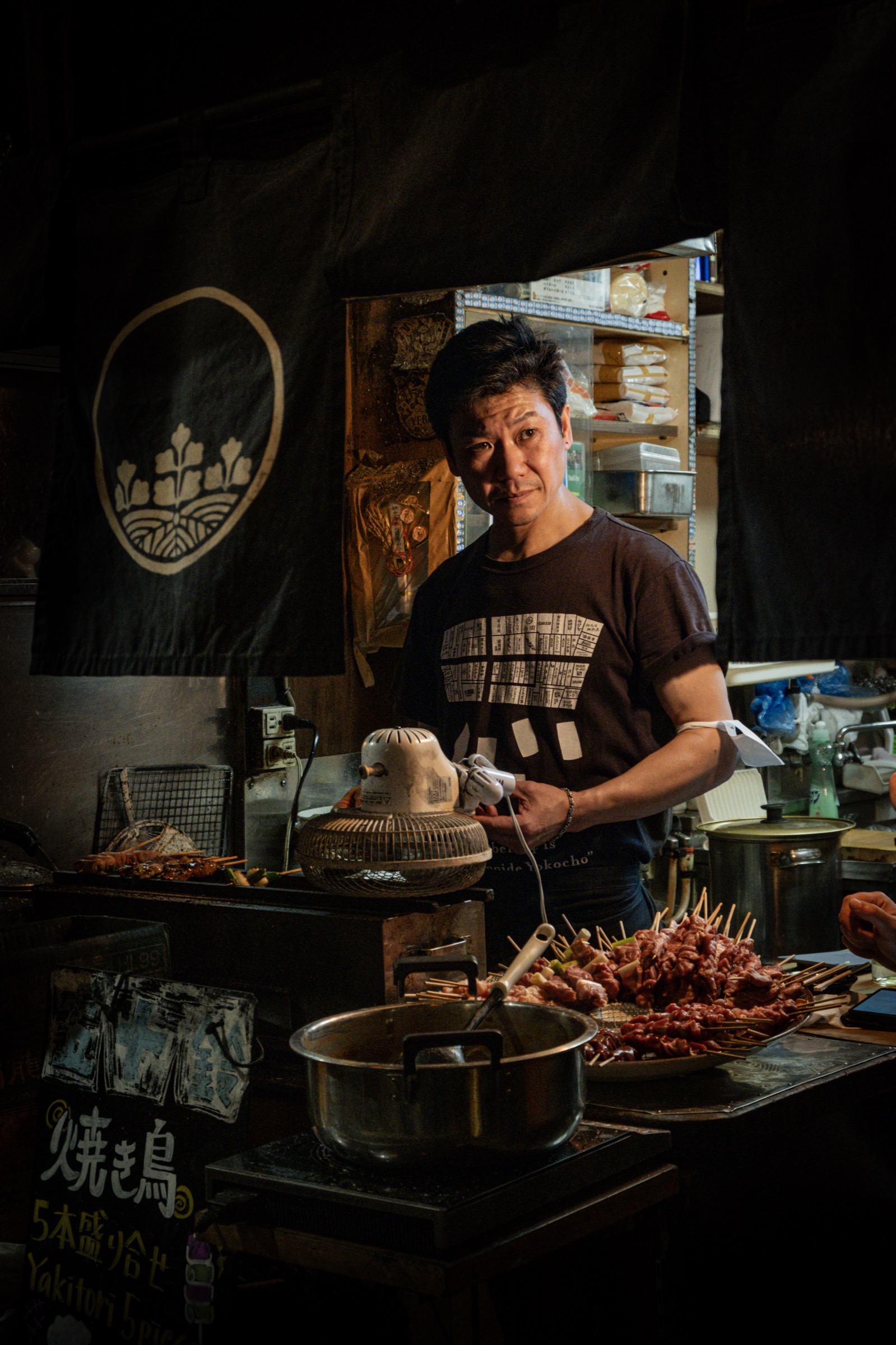 A woman grills yakitori skewers over charcoal behind a dark noren curtain with a family-crest emblem