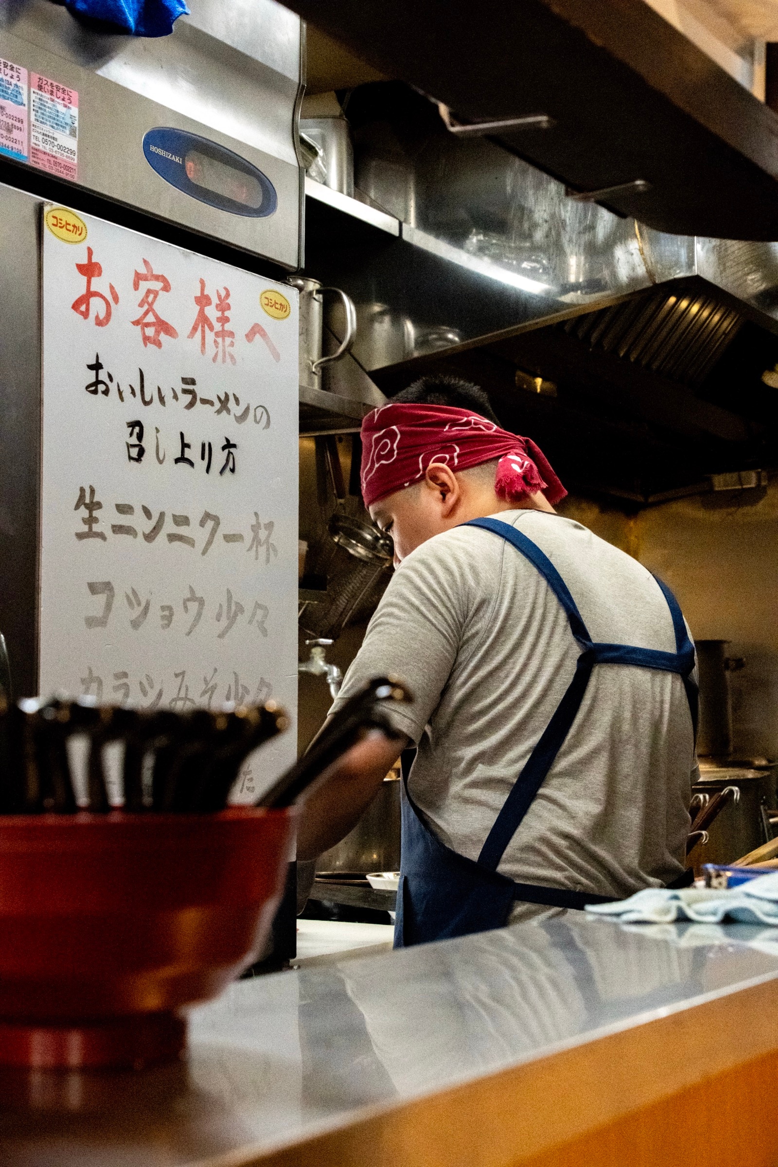 A ramen cook in a red bandana and apron works at the counter beside a hand-painted sign with serving instructions
