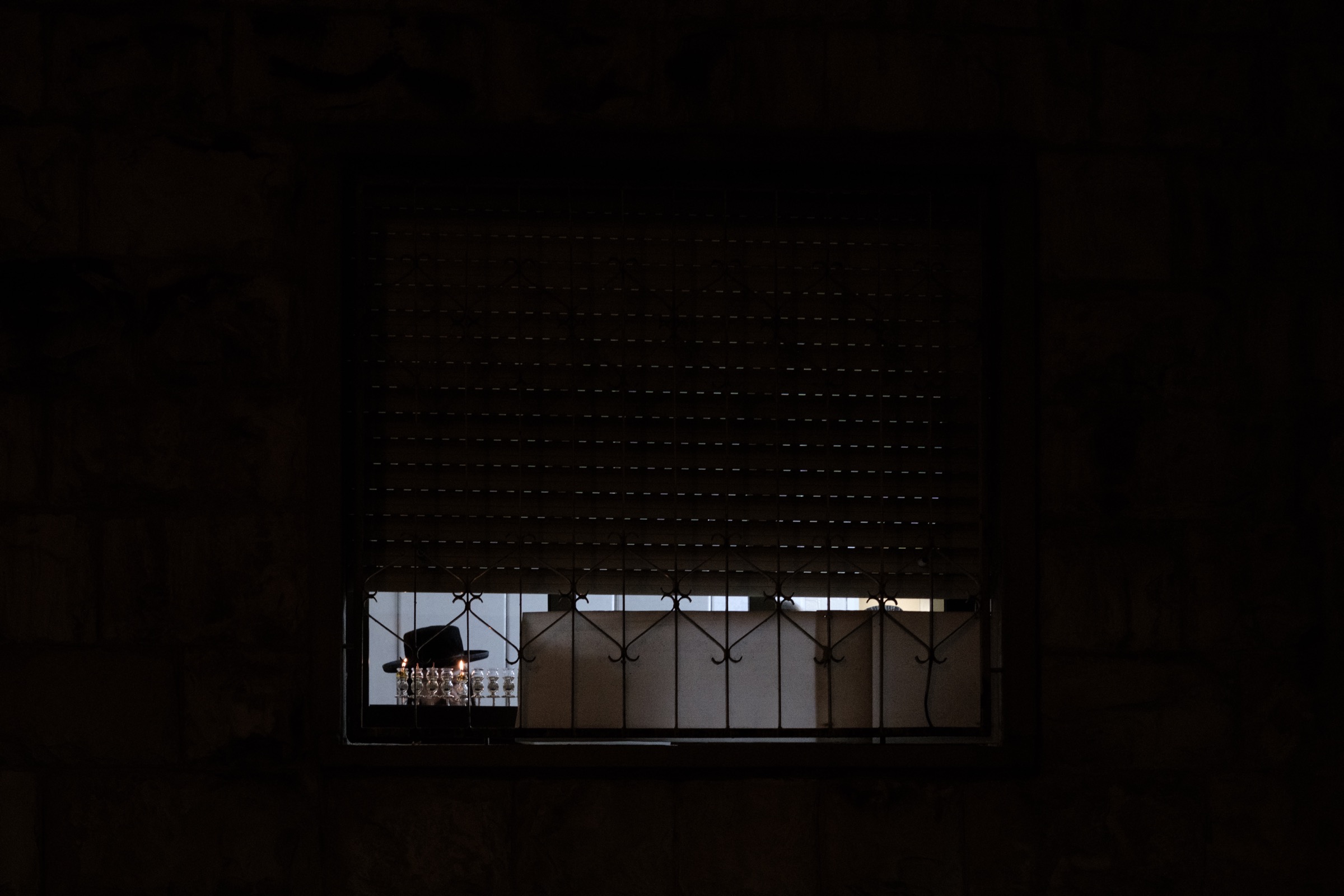 A lit menorah seen through a window with half-closed blinds and iron bars, the top of a black hat just visible below