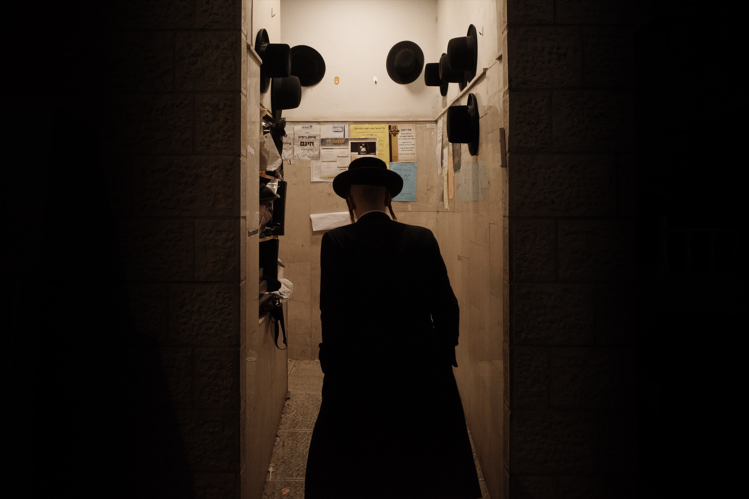 A man in a black hat walks into a narrow hallway entrance with black hats hanging on hooks along both walls