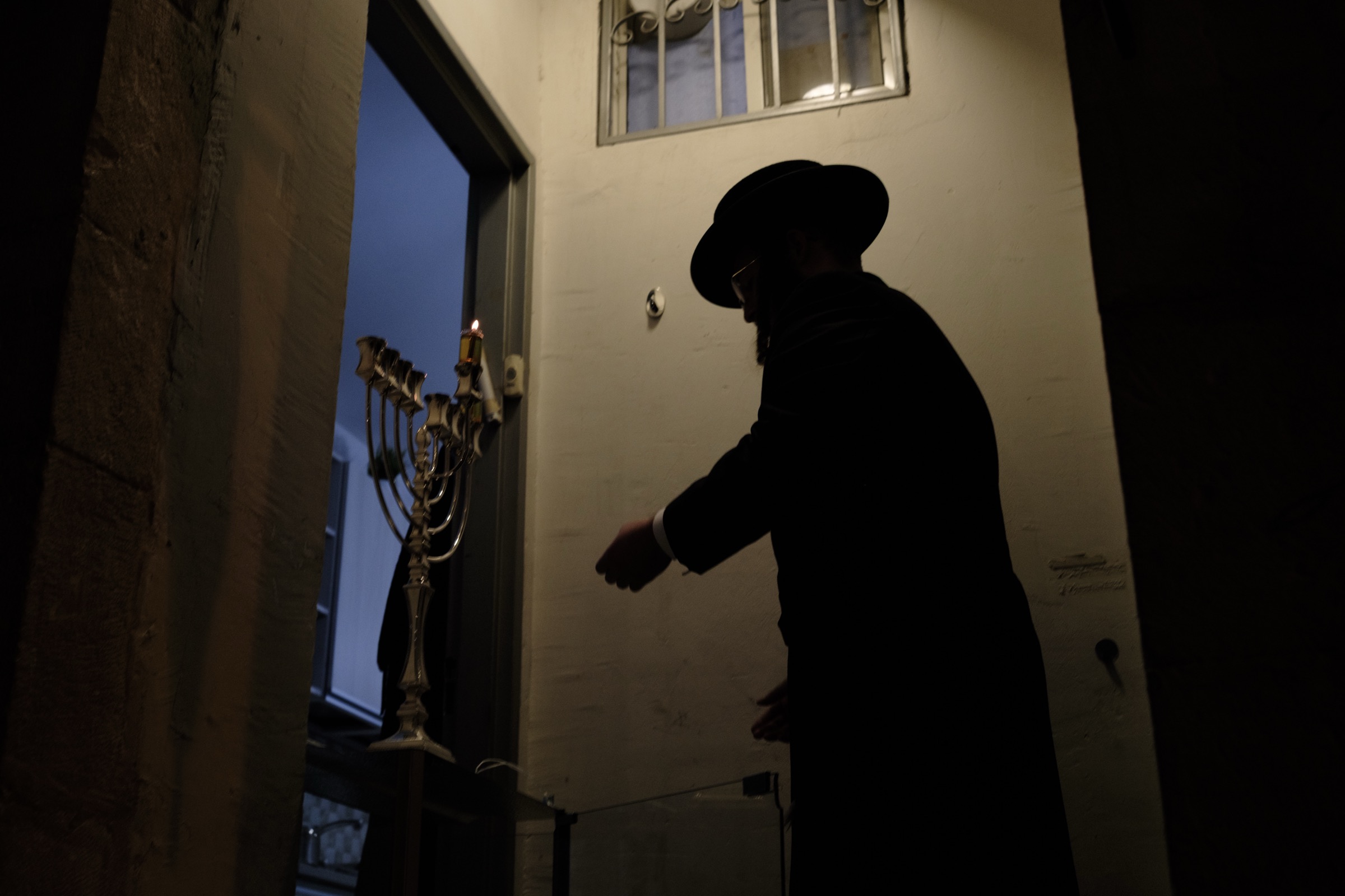 Silhouette of a man in a black coat and hat standing beside a silver menorah with a single lit candle in a doorway
