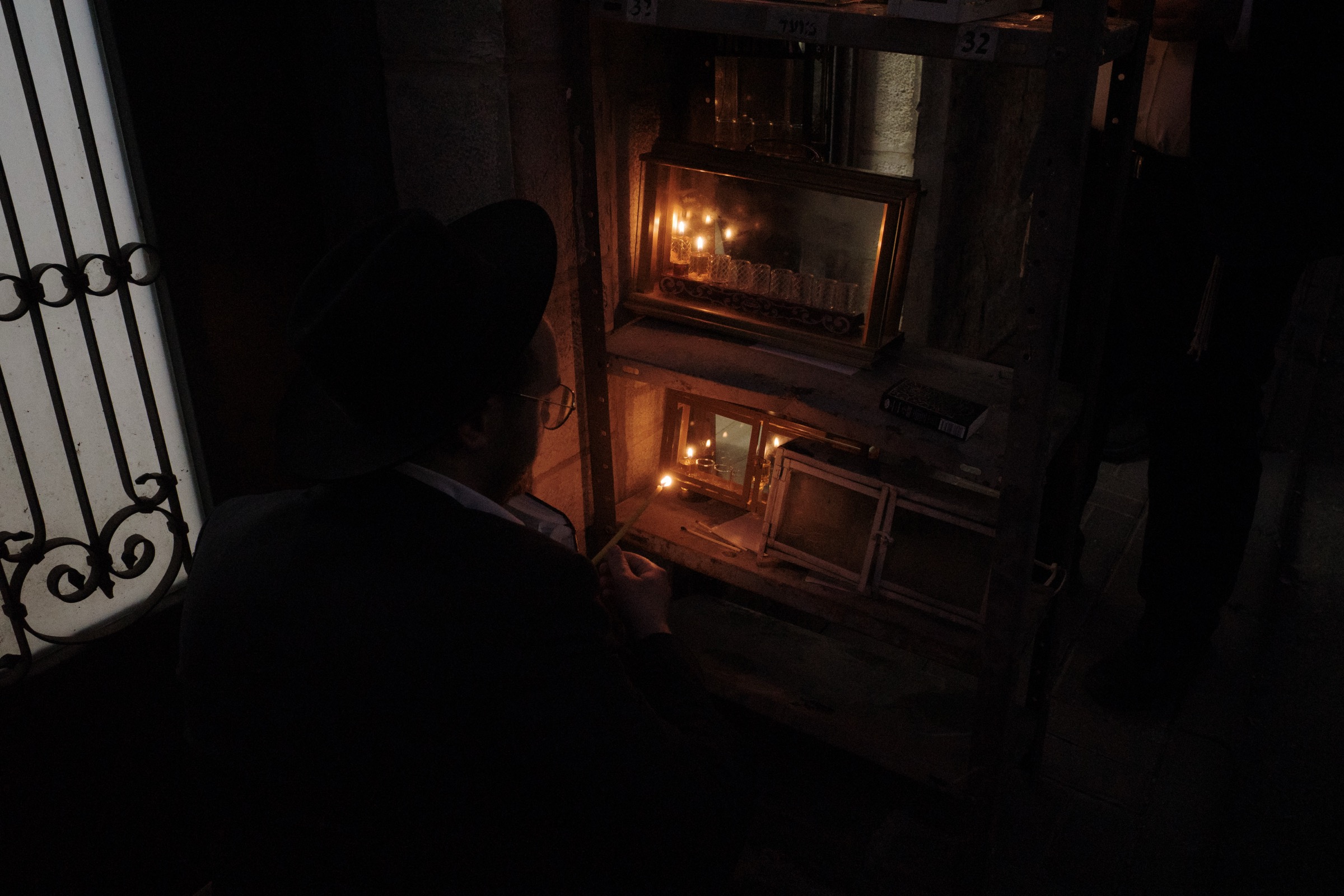 A man in a black hat crouches to light Hanukkah candles in glass-enclosed menorahs outside a doorway